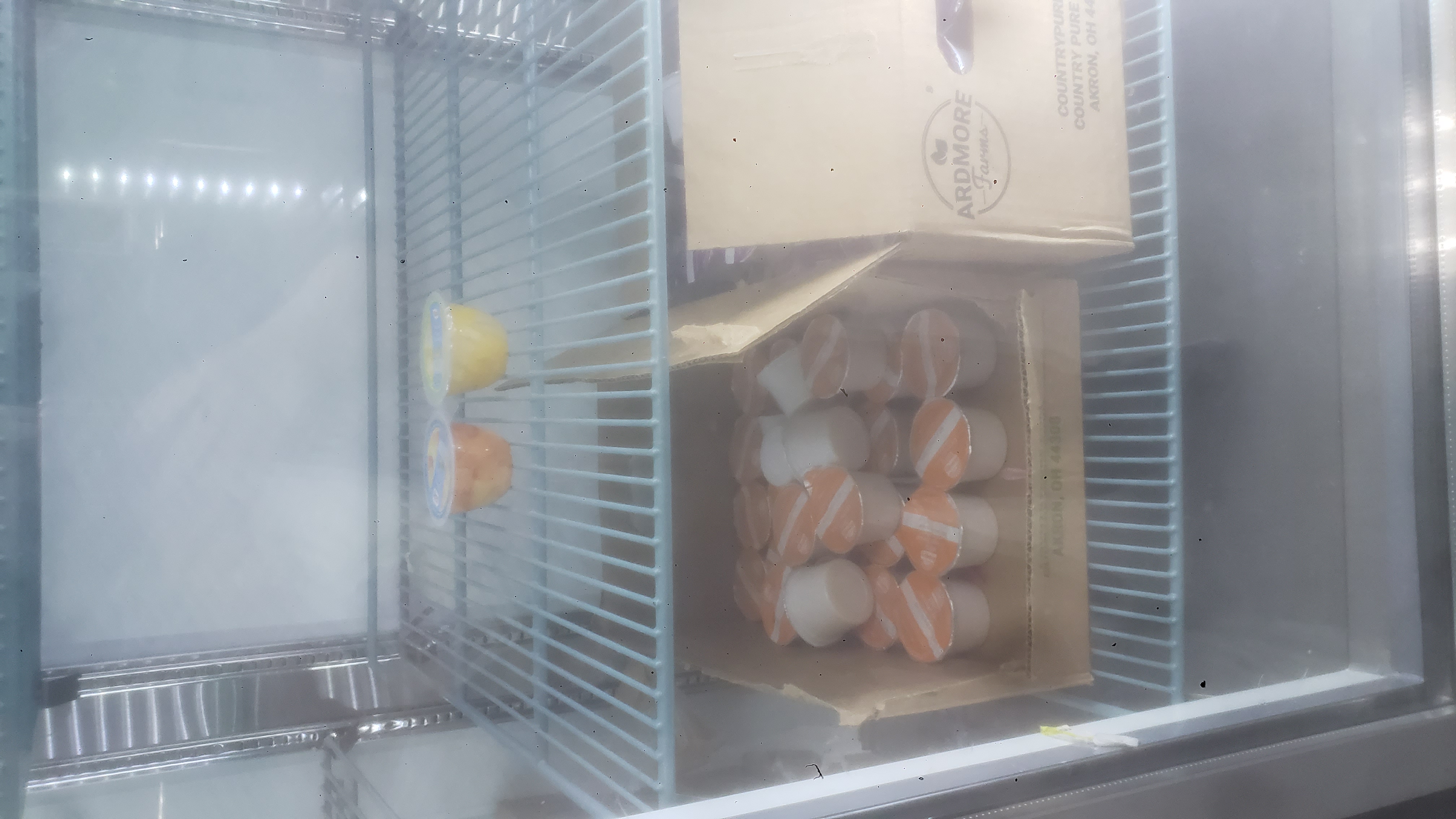 Two cups of fruits on one shelf and boxes full of mysterious cups on the bottom of a fridge.